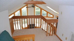 Una habitación con techo inclinado, vigas de madera, una pequeña escalera y vistas a la vegetación.