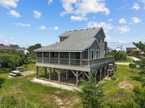 Une maison située sur la côte, dotée d’une terrasse en bois et d’un balcon, entourée de verdure et surplombant un ciel dégagé.