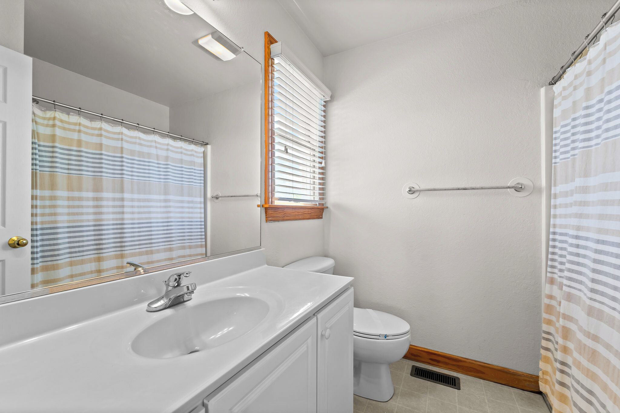 A bathroom with a white sink, mirror, and toilet, a window with blinds, and a shower curtain.