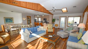 A spacious living room with a blue sofa, striped armchair, and a wooden coffee table. There is a dining area with a table and chairs, and a kitchen in the background. - Awesome 'D' has a hot tub for your family vacation entertainment (South Nags Head)