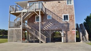 A two-story building with a wooden exterior, a balcony on the upper floor, and a staircase leading to a lower level. - Awesome 'D' has a hot tub for your family vacation entertainment (South Nags Head)