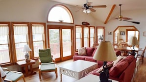 A spacious living room with a red sofa, green armchair, and white coffee table. There are large windows with curtains, a ceiling fan, and a dining area in the background. - When it is time to Set Back & Relax, you have found the vacation rental home to do just that. (Corolla)