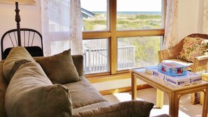 A cozy living room with a large window, a sofa, a chair, and a table with a box of books. - Dreamcatcher is a well-equipped vacation home perfect for a terrific vacation experience. (Corolla)
