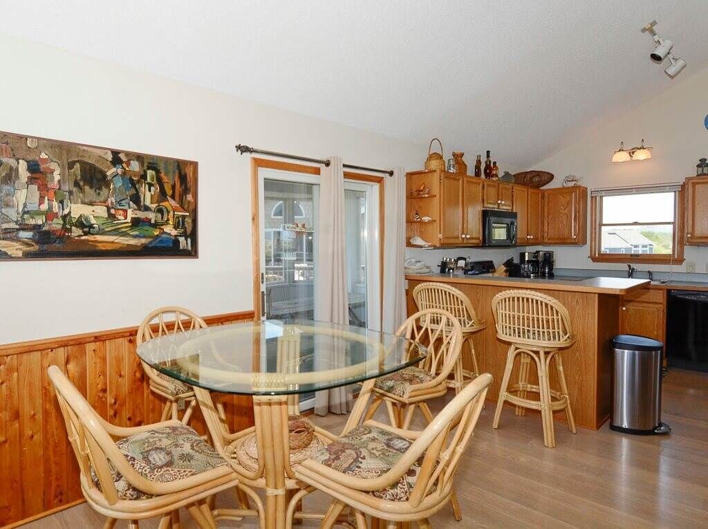 A modern kitchen with wooden cabinets, a glass dining table, wicker chairs, and a painting on the wall.