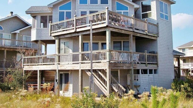 A multi-story house with a balcony on the second floor, a covered porch on the ground floor, and a chimney on the roof. - Dreamcatcher is a well-equipped vacation home perfect for a terrific vacation experience. (Corolla)