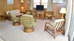 A cozy room with a wooden paneled wall, a television on a wooden cabinet, wicker furniture, and a window with curtains. - Just across the street from the beach, “Pope” is perfect for a small family. (South Nags Head)