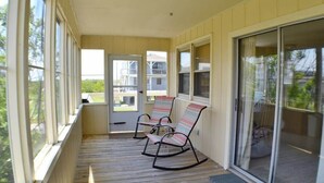 A covered porch with two rocking chairs, a sliding glass door, and a view of a residential area. - Just across the street from the beach, “Pope” is perfect for a small family. (South Nags Head)