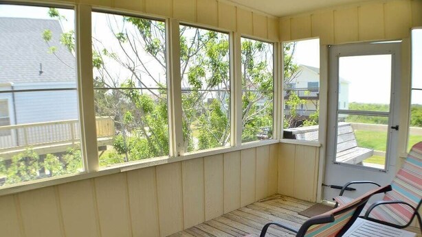 A covered porch with wooden flooring, two striped chairs, and a door leading outside. - Just across the street from the beach, “Pope” is perfect for a small family. (South Nags Head)
