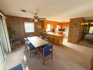 A kitchen with wooden cabinets, a central island, and a dining area with blue chairs. - Great home on the OBX! (Kill Devil Hills)