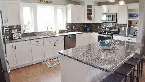 A modern kitchen with white cabinets, a granite countertop, and a central island with bar stools. - Charming Outer Banks Home (Kill Devil Hills)