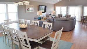 A spacious living area with a dark wooden dining table surrounded by white chairs, a grey sectional sofa, and a ceiling fan. - Charming Outer Banks Home (Kill Devil Hills)