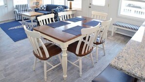 Un coin repas avec une table en bois et des chaises blanches, un tapis bleu et un banc près de la fenêtre.