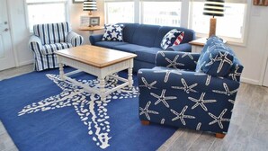 Un salon avec un canapé bleu, un fauteuil à rayures et un pouf, une table basse en bois et un tapis orné de motifs d’étoiles de mer.