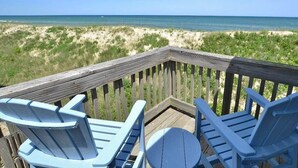 Una terraza de madera con sillas Adirondack azules y una mesa redonda, con vista a una playa con dunas y el océano.