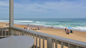 Une vue sur la plage depuis un espace abrité, équipé d’une table et de chaises, d’où l’on peut admirer l’océan et les personnes se promenant sur la plage.