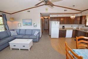 A living room with a sectional sofa, a white coffee table, and a dining area with a wooden table and chairs. A kitchen with wooden cabinets and a refrigerator is visible in the background. - Book into Brooks and you will not be disappointed! (Nags Head)
