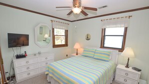 Un dormitorio con cama, mesitas de noche, cómoda, ventilador de techo y televisor.