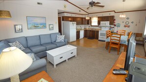 Una sala de estar con sofá, mesa de centro y lámpara. Hay una zona de cocina con refrigerador, microondas y mesa de comedor con sillas.