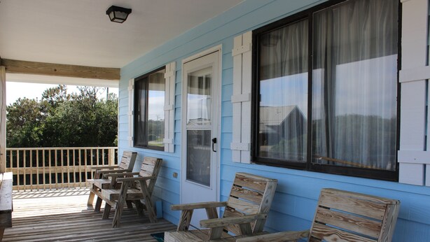 Una terraza de madera con varias sillas y una mesa, una puerta y ventanas con cortinas.