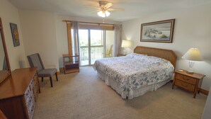 A bedroom with a large bed, a chair, a dresser, a nightstand with a lamp, and a ceiling fan.
