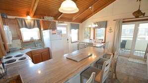 A kitchen with wooden cabinets, a central island, a stove, and a dining area with chairs and a table. - Listen to the waves break and look for dolphins right from your screened-in porch! (South Nags Head)