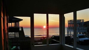 A view from a room with large windows overlooking a sunset over the ocean, with a deck and outdoor seating area visible. - Listen to the waves break and look for dolphins right from your screened-in porch! (South Nags Head)