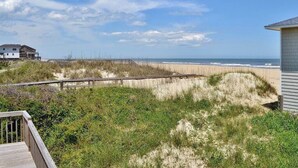 A beach view with a wooden deck, a sandy dune, and a building in the background. - Listen to the waves break and look for dolphins right from your screened-in porch! (South Nags Head)