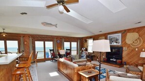 A spacious living room with a wooden floor, a large sofa, a coffee table, and a television. There is a bar area with stools and a view of the ocean. - This charming beach house is the perfect year-round retreat. (South Nags Head)