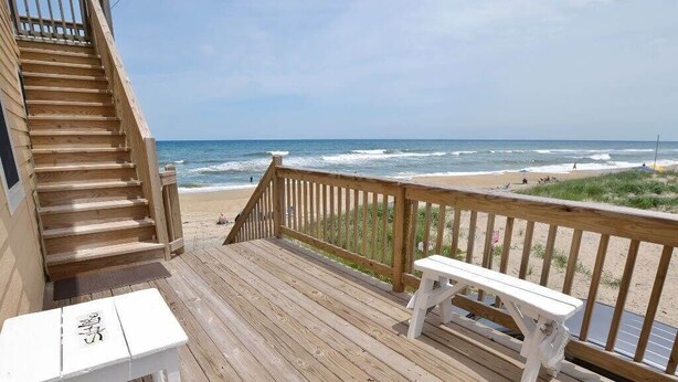 A wooden deck with a bench and a table, overlooking a beach with waves and a clear sky. - This charming beach house is the perfect year-round retreat. (South Nags Head)