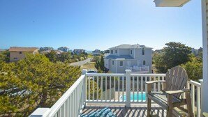 A deck with a pool, a chair, and a view of a large house and beach.