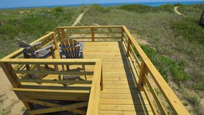 Eine Holzterrasse mit zwei Adirondack-Stühlen und einem Tisch, mit Blick auf einen Strand und einen klaren Ozean.