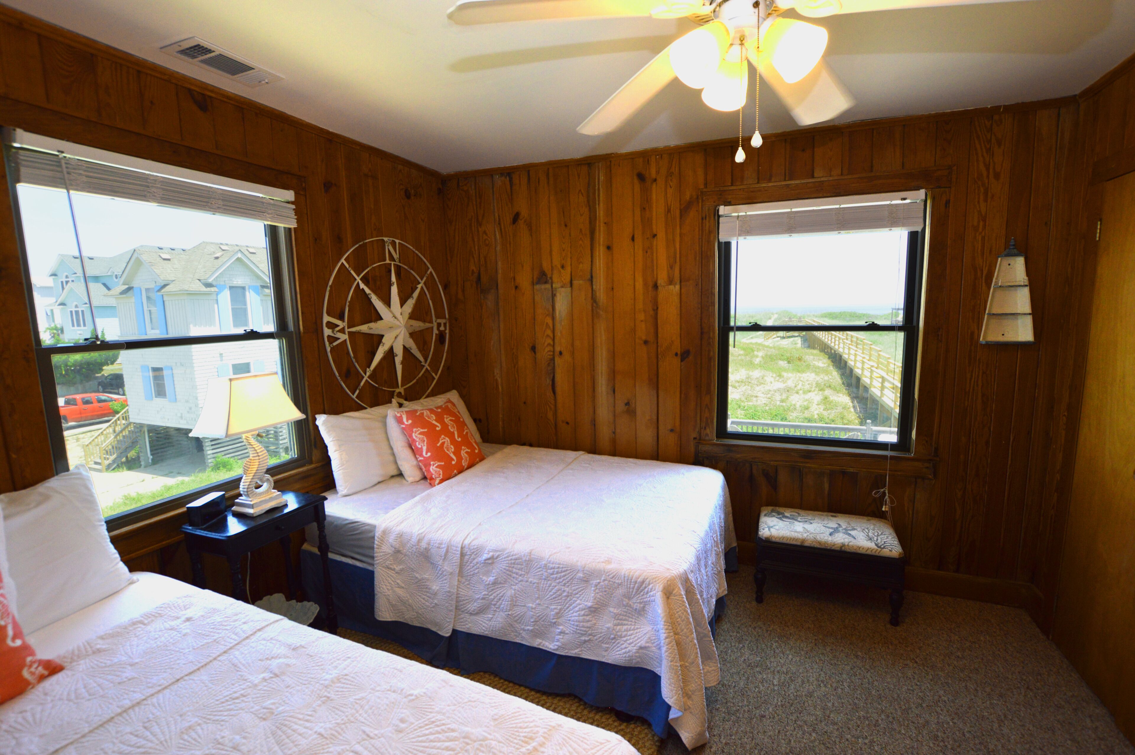 A room with wooden paneling, two beds, a ceiling fan, a compass wall decoration, and large windows with views of houses.