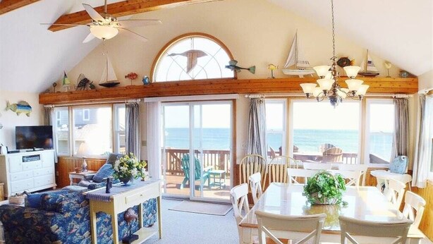 Un vaste espace de vie doté d’une grande fenêtre donnant sur la mer, de poutres apparentes en bois au plafond, d’une table à manger avec des chaises et d’un canapé à motifs floraux.