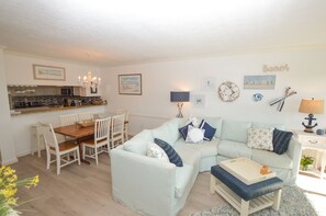 A bright living room with a light blue sofa, a wooden dining table with chairs, and a kitchen area in the background.