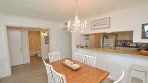 A dining area with a wooden table and white chairs, a chandelier, and a kitchen in the background. - Great condo in Duck, NC (Duck)