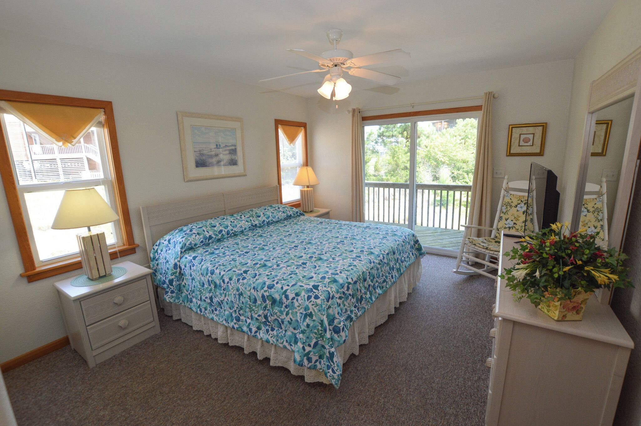 A bedroom with a bed, a nightstand, a lamp, a dresser, a mirror, and a window with a view of greenery.