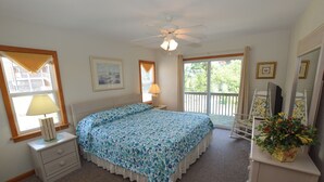 A bedroom with a bed, a nightstand, a lamp, a dresser, a mirror, and a window with a view of greenery. - Enjoy ocean views from this beautiful home, nestled in a quiet soundside community. (Nags Head)