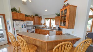 A kitchen with wooden cabinets, a central island with bar stools, a refrigerator, microwave, and dishwasher. - Enjoy ocean views from this beautiful home, nestled in a quiet soundside community. (Nags Head)