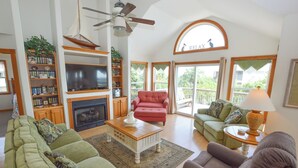 A spacious living room with a fireplace, a television, and a bookshelf. There are multiple seating areas with green and brown sofas. - Enjoy ocean views from this beautiful home, nestled in a quiet soundside community. (Nags Head)