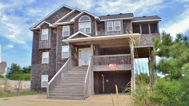 A two-story house with a front porch, a staircase leading to the entrance, and a garage. - Enjoy ocean views from this beautiful home, nestled in a quiet soundside community. (Nags Head)
