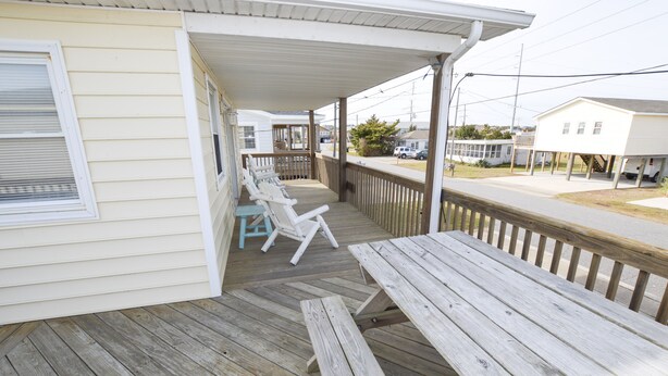 A covered porch with wooden benches and a picnic table, overlooking a street with houses and parked cars. - Pet Friendly on Outer Banks. (Kill Devil Hills)