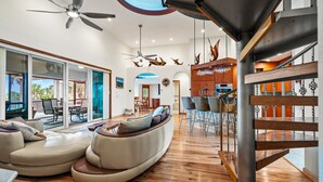 A spacious living area with a curved sofa, a dining area with chairs, a kitchen with bar seating, and a staircase with wooden railings. - Stella Marris- N. Captiva Island | Luxury Beachfront Retreat (Lee County)