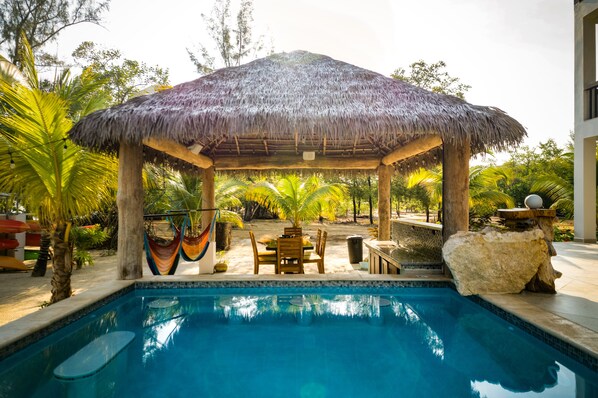 A pool area with a thatched-roof gazebo, wooden furniture, and a stone feature.