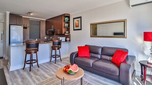 A modern living room with a brown leather sofa, two bar stools, a round coffee table with fruit, and a kitchen area with a refrigerator, stove, and bar counter. - Quayside 1206by CTHA (Cape Town)