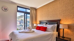 A hotel room with a large bed, a nightstand with a lamp, a clock on the wall, and a window offering a view of buildings and hills. - Quayside 1206by CTHA (Cape Town)