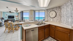 A modern kitchen with a dishwasher, wooden cabinets, and a large window overlooking the ocean. - AquaVista by Book That Condo (Panama City Beach)