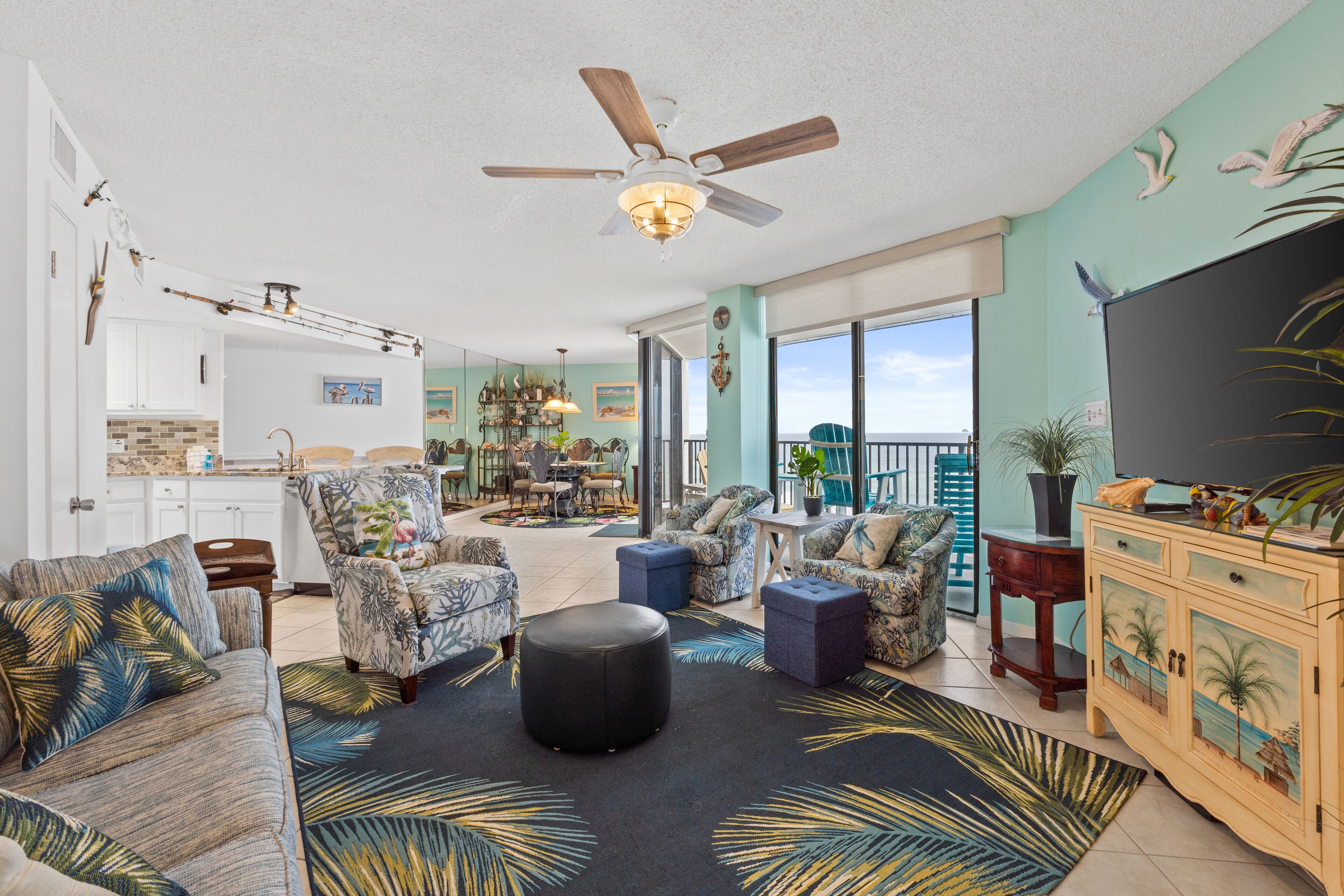 A spacious living room with a ceiling fan, a large TV, and a balcony with a view.