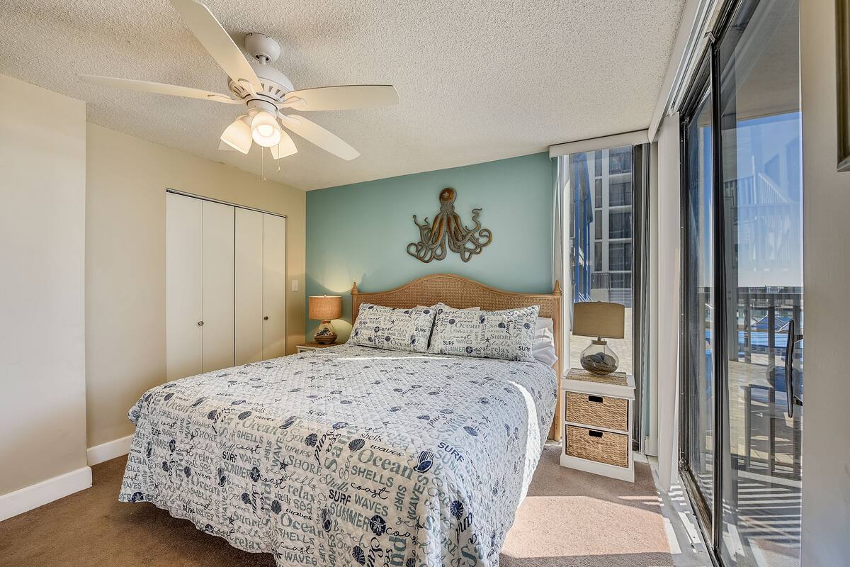 A bedroom with a bed, a ceiling fan, a wall-mounted lamp, a decorative octopus wall art, and a view of the outside through a sliding glass door.