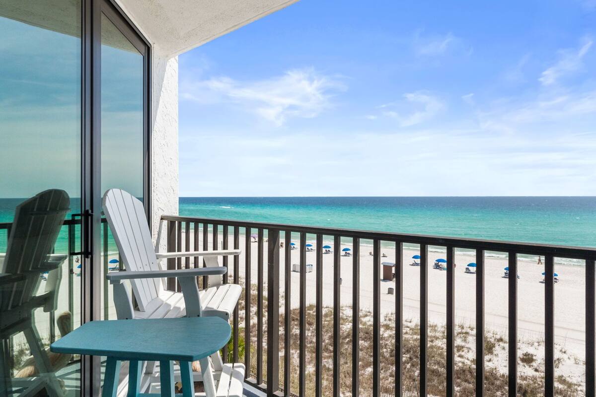 A balcony with white Adirondack chairs and a table, overlooking a beach with umbrellas and people.