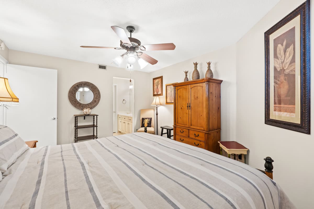 A bedroom with a bed, a ceiling fan, a wooden cabinet, a mirror, and a framed picture on the wall.